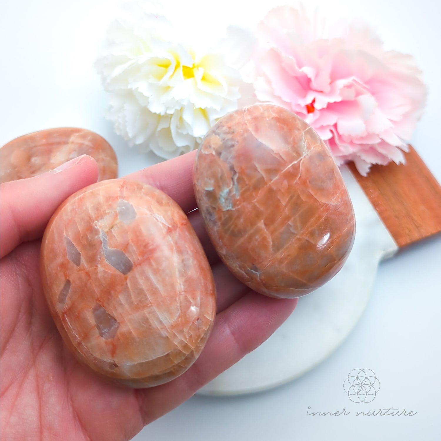 Hand holding 2 pink stone swith floral background. Pink/Peach moonstone palm stone available at Inner Nurture, an online crystal shop in Australia. 