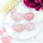 Pink heart-shaped objects on a white marble surface with flowers. Rose Quartz hearts available at Inner Nurture, an online crystal shop in Australia.