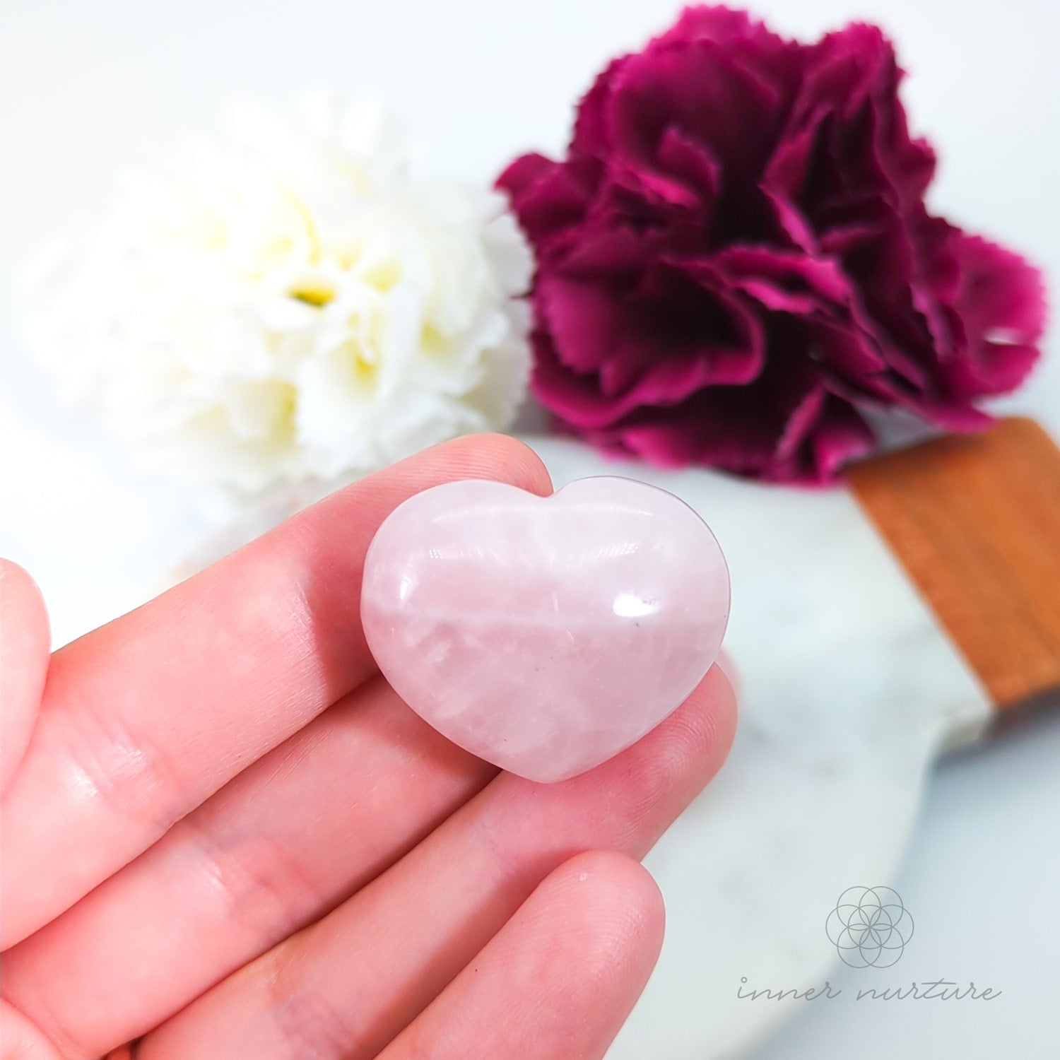 Heart-shaped rose quartz crystal held in a hand with flowers in the background. Rose Quartz hearts available at Inner Nurture, an online crystal shop in Australia.