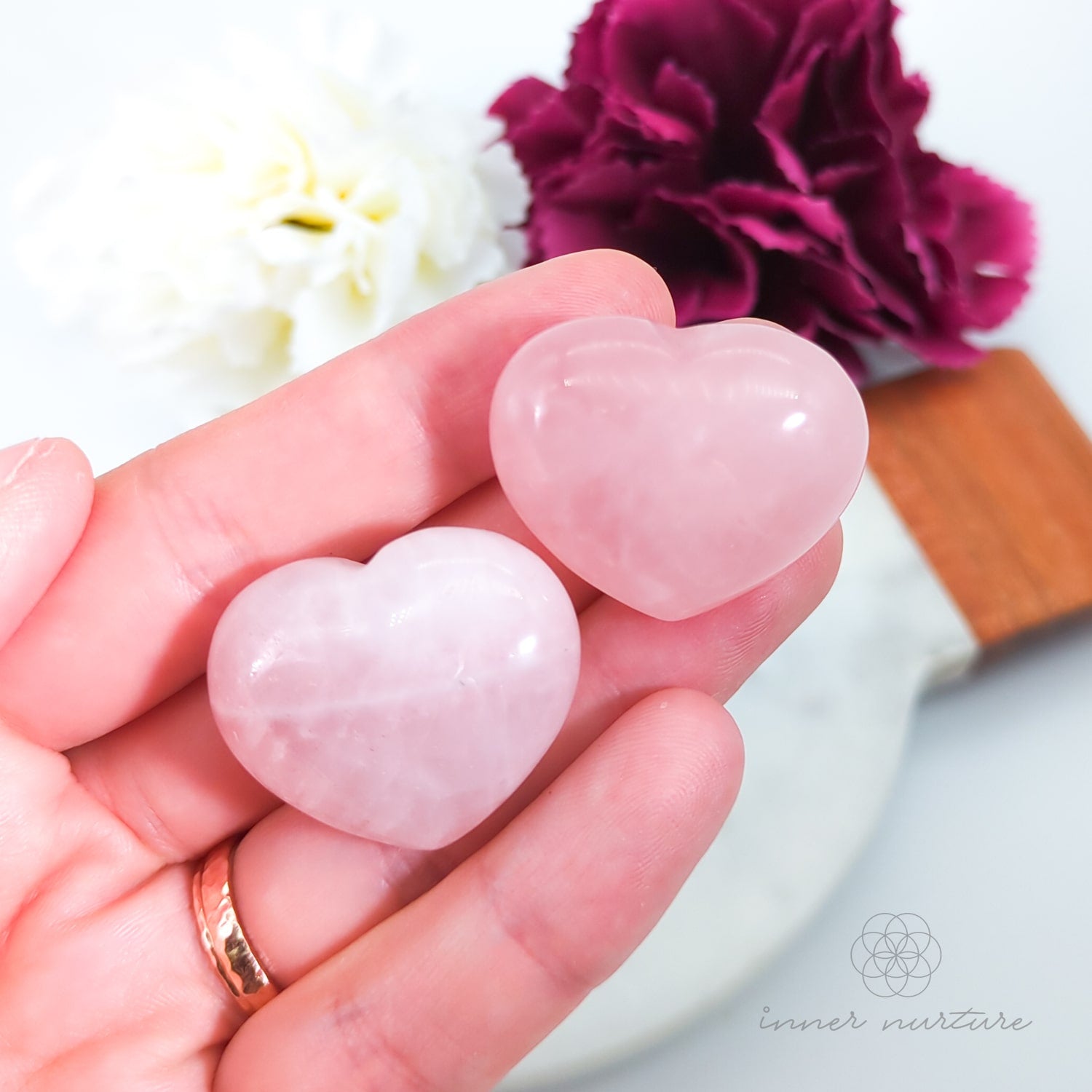 Heart-shaped pink stones held in a hand with flowers in the background.  Rose Quartz hearts available at Inner Nurture, an online crystal shop in Australia.