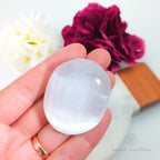 Clear crystal stone held in a hand with flowers and a wooden tool in the background.  Selenite palm stone available at Inner Nurture, an online crystal shop in Australia.