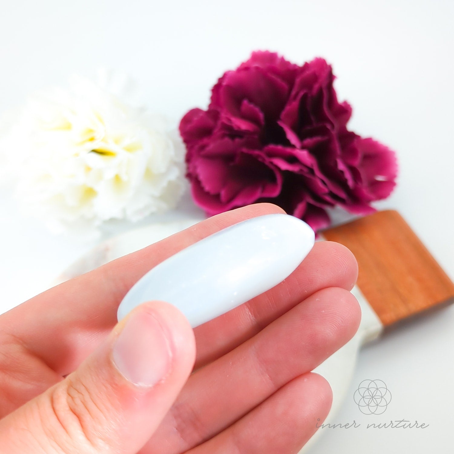 Hand holding a white object with flowers and a wooden block in the background. Selenite palm stone available at Inner Nurture, an online crystal shop in Australia.