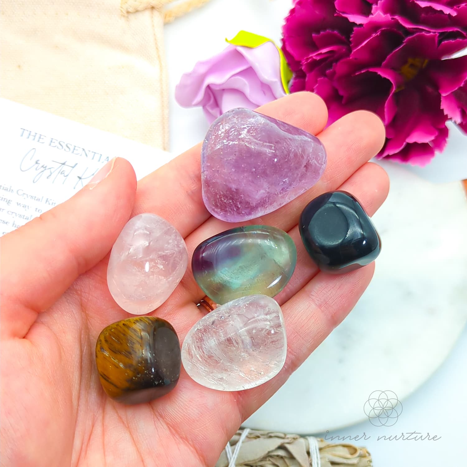 Hand holding a collection of colorful crystals with flowers in the background. Essentials Crystal Kit, available on line in Australia at Inner Nurture