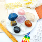 Set of colorful crystals on a white plate with flowers and a candle in the background. Chakra Balancing Crystal Kit, available on line in Australia at Inner Nurture