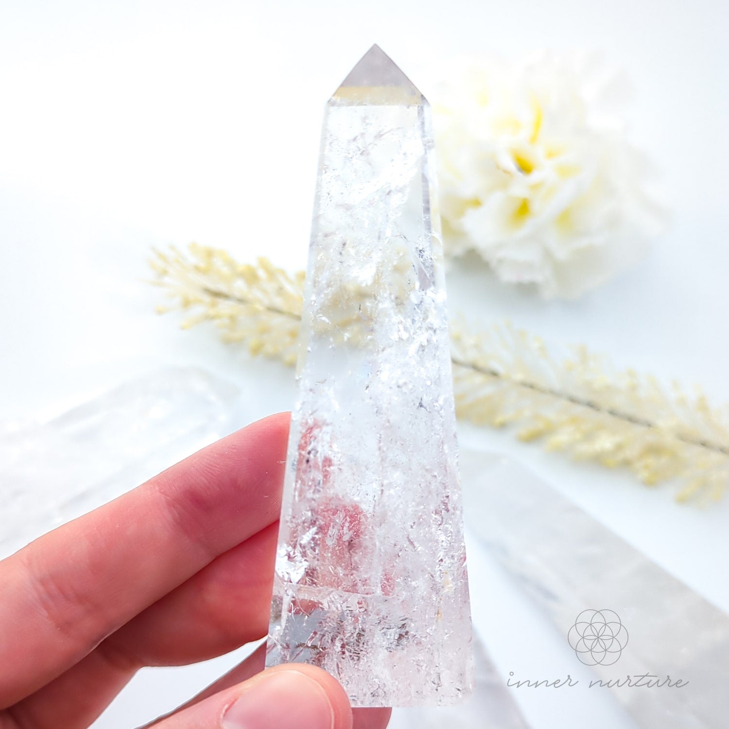 Clear crystal tower held by a hand with a soft white background and flowers. Clear Quartz Tower for Clarity & Energy | Crystals Australia Inner Nurture