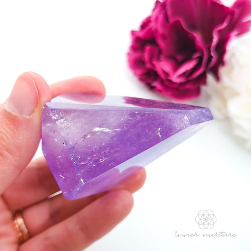 Hand holding a purple crystal with pink flowers in the background. Amethyst Free Form #3 for Calm & Clarity | Crystals Australia