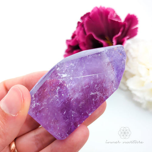 Hand holding a purple crystal with a pink flower in the background. Amethyst Free Form #3 for Calm & Clarity | Crystals Australia