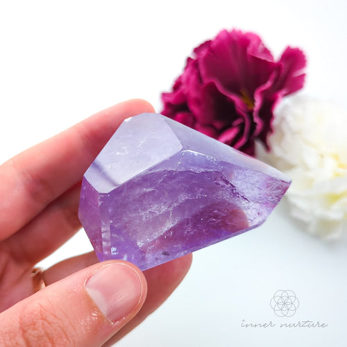 Hand holding a purple crystal with flowers in the background. Amethyst Free Form #3 for Calm & Clarity | Crystals Australia