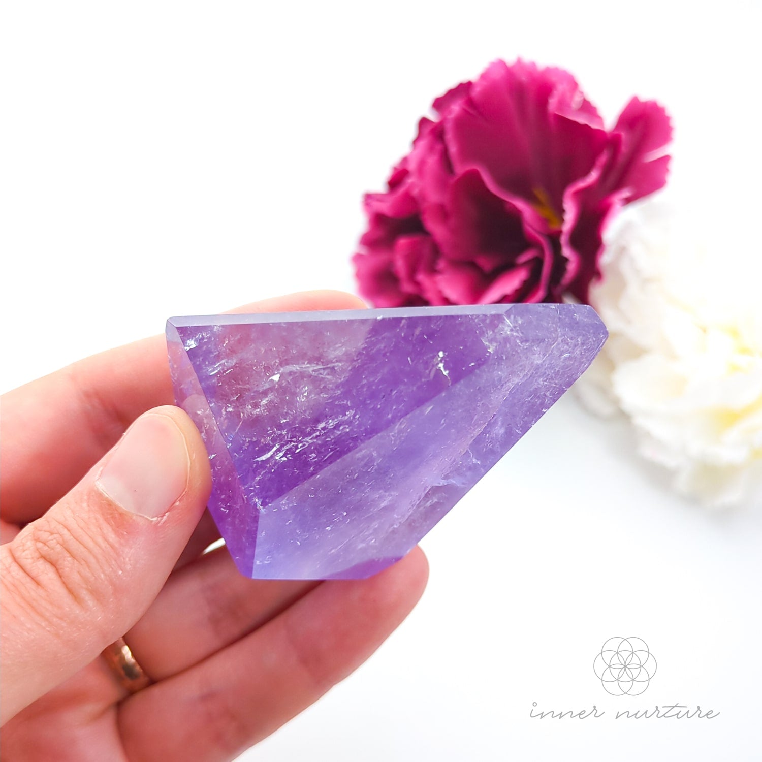 Hand holding a purple crystal with a pink flower in the background. Amethyst Free Form #3 for Calm & Clarity | Crystals Australia