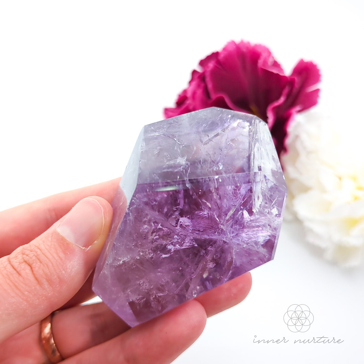 Hand holding a purple crystal with a white flower and pink flower in the background. Amethyst Free Form #2 for Calm & Clarity | Crystals Australia