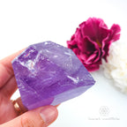 Hand holding a purple crystal with pink flowers in the background. Amethyst Free Form #1 for Calm & Clarity | Crystals Australia