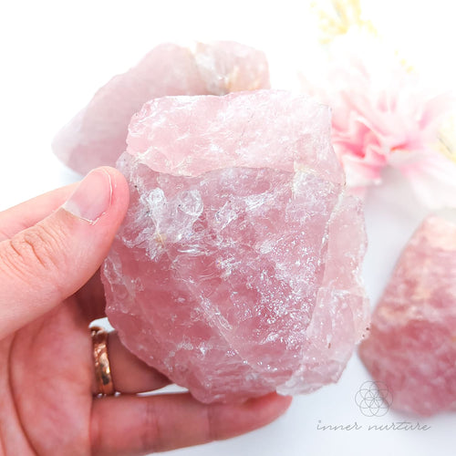 Hand holding a pink crystal rock with a soft focus floral background. Rose Quartz raw crystal available online at Inner Nurture Crystals Australia