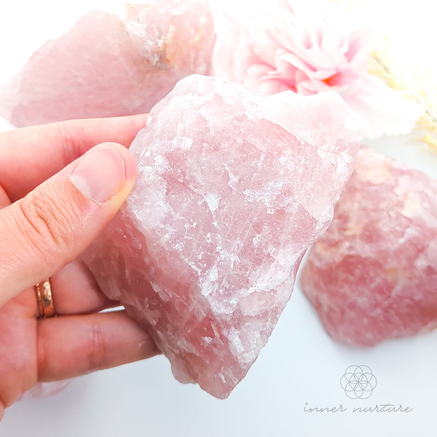 Hand holding a pink crystal with more crystals in the background on a white surface. Rose Quartz raw crystal available online at Inner Nurture Crystals Australia