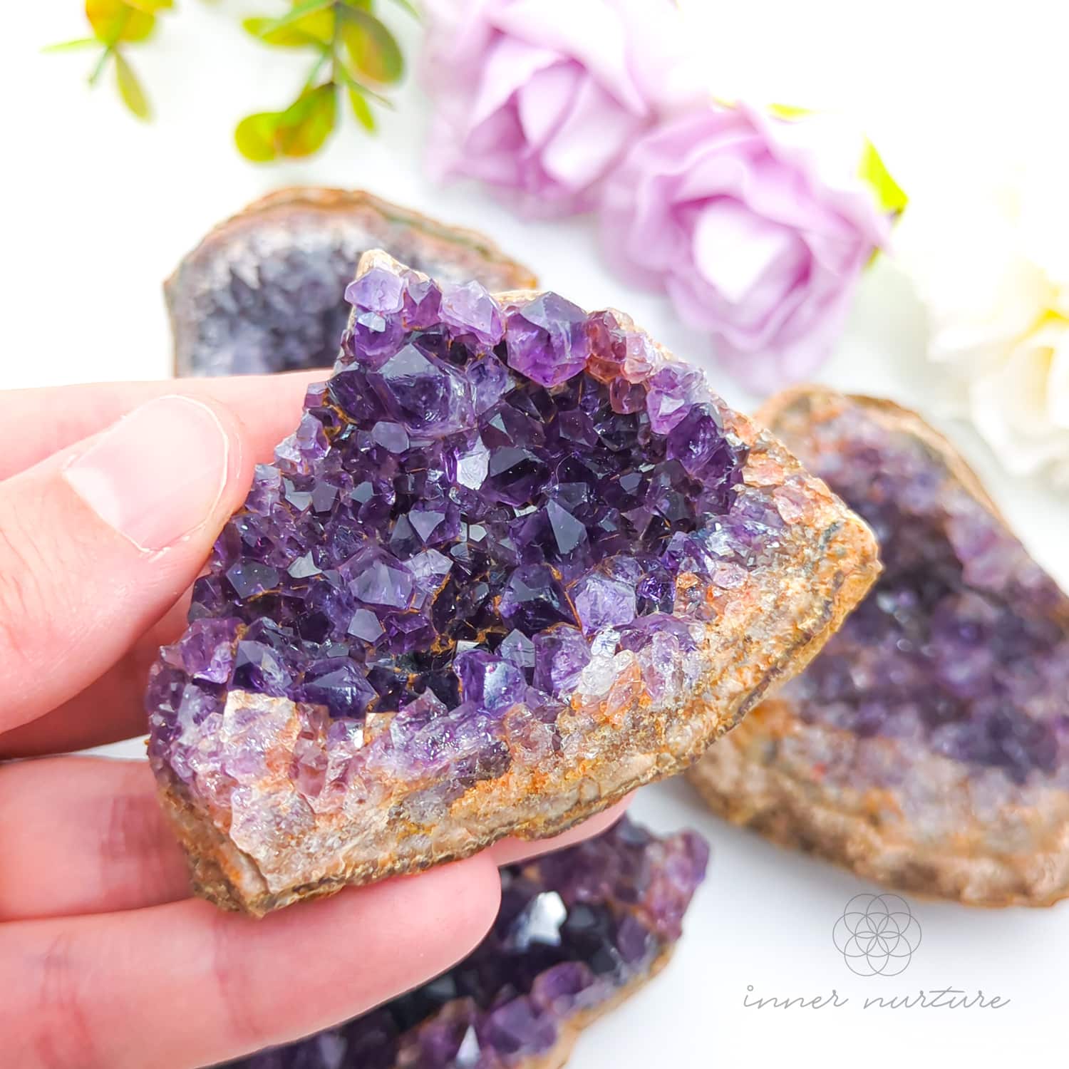 Hand holding a purple crystal rock with more crystals and flowers in the background. Amethyst crystal cluster available online at Inner Nurture Crystals Australia