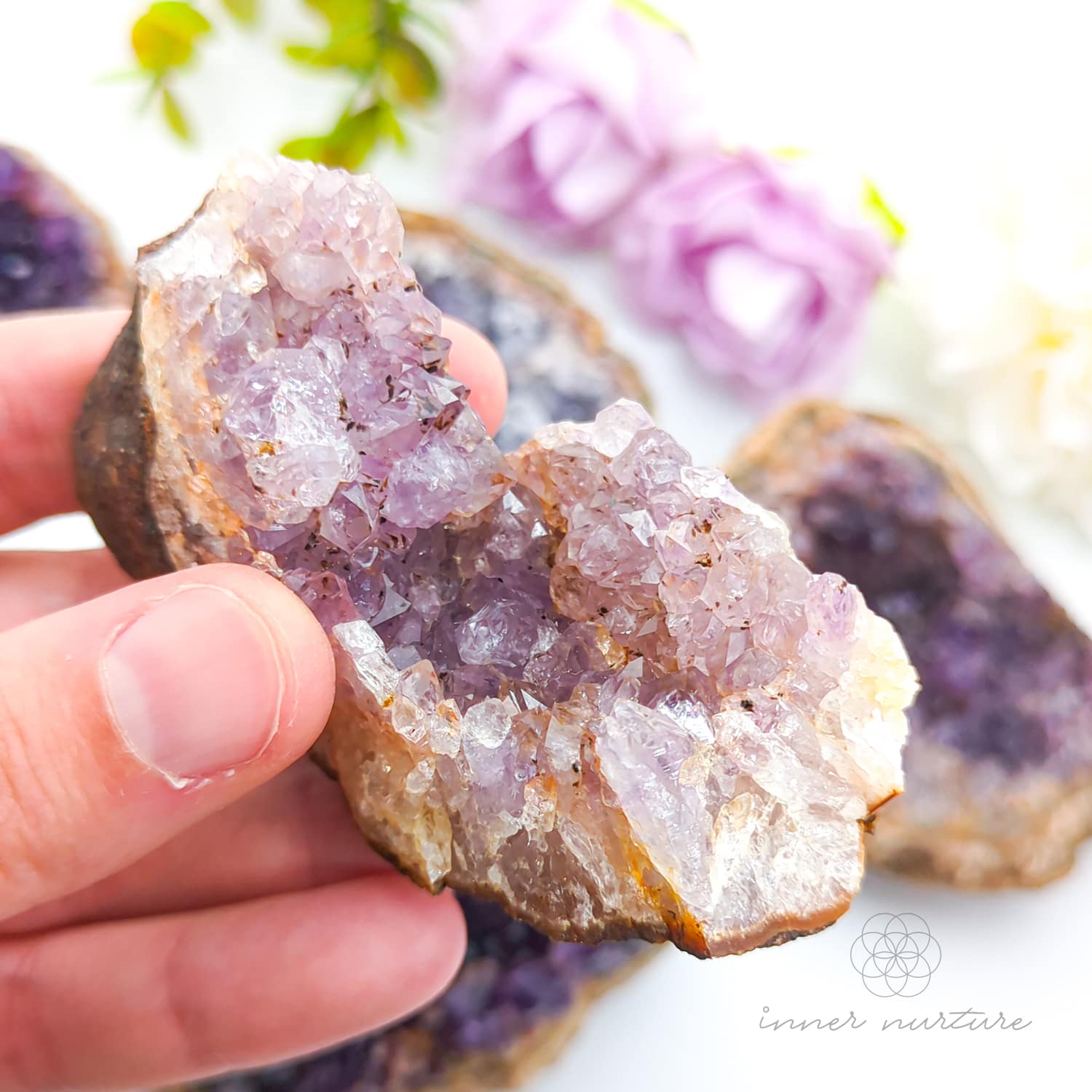 Hand holding a purple crystal rock with blurred flowers in the background. Amethyst crystal cluster available online at Inner Nurture Crystals Australia