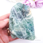 Hand holding a large green crystal with flowers in the background. Rainbow Fluorite available at Inner Nurture crystals australia. 