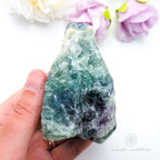 Hand holding a large green crystal with flowers in the background. Rainbow Fluorite available at Inner Nurture crystals australia. 