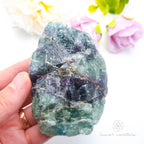 Hand holding a large crystal with colorful flowers in the background. Rainbow fluorite available at Inner Nurture crystals australia