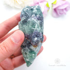Hand holding a large crystal with colorful flowers in the background. Rainbow fluorite available at Inner Nurture crystals australia