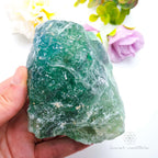 Hand holding a large green crystal with flowers in the background. Rainbow Fluorite available at Inner Nurture crystals australia