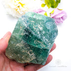 Hand holding a large green crystal with flowers in the background. Rainbow Fluorite available at Inner Nurture crystals australia