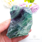Rainbow fluorite specimen held in hand with purple flowers in background. Available at Inner Nurture crystals in Australia