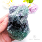 Rainbow fluorite specimen held in hand with purple flowers in background. Available at Inner Nurture crystals in Australia