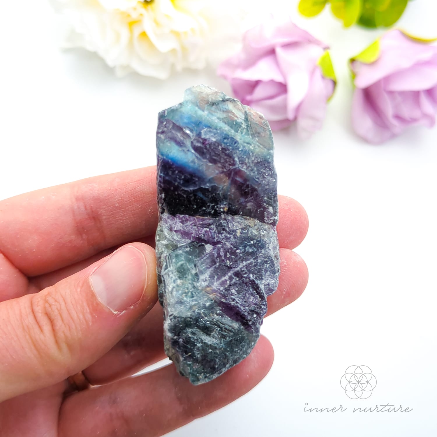 Hand holding a multicolored crystal with flowers in the background. rainbow fluorite available at Inner Nurture Crystals in australia