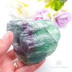 Raw fluorite crystal held in a hand with a blurred floral background. rainbow fluorite available at Inner Nurture crystals in Australia