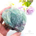 Raw fluorite crystal held in a hand with a blurred floral background. rainbow fluorite available at Inner Nurture crystals in Australia
