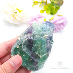 Hand holding a green crystal with flowers in the background. Rainbow Fluorite available at Inner Nurture crystals in australia