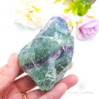 Hand holding a green crystal with flowers in the background. Rainbow Fluorite available at Inner Nurture crystals in australia