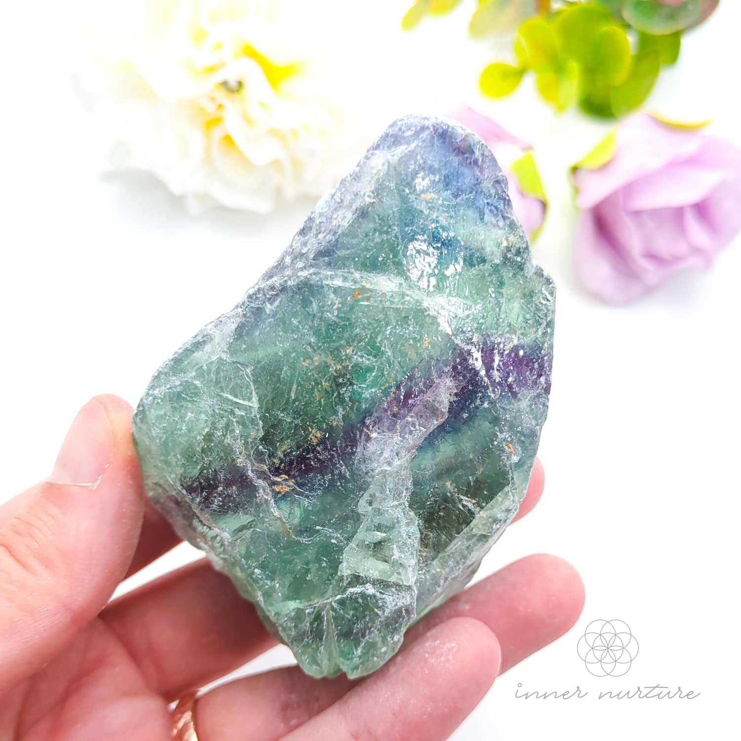 Hand holding a green crystal with flowers in the background. Rainbow Fluorite available at Inner Nurture crystals in australia