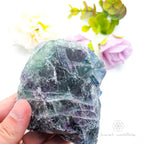 Raw fluorite crystal held by a hand with a soft focus floral background .Rainbow Fluorite available at Inner Nurture crystals in Australia