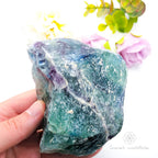 Hand holding a large crystal rock with a blurred floral background. Rainbow Fluorite available at Inner Nurture crystals in Australia