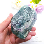 Hand holding a large crystal rock with a blurred floral background. Rainbow Fluorite available at Inner Nurture crystals in Australia
