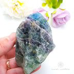Raw crystal held in a hand with flowers in the background. Rainbow Fluorite available at Inner Nurture crystals in Australia