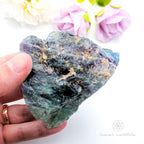 Raw crystal held in a hand with flowers in the background. Rainbow Fluorite available at Inner Nurture crystals in Australia