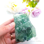 Hand holding a green crystal with flowers in the background. Rainbow Fluorite available at Inner Nurture crystals australia