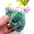 Hand holding a green crystal with flowers in the background. Rainbow Fluorite available at Inner Nurture crystals australia