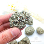 Hand holding a pyrite crystal with more crystals on a white background. Pyrite crystal cluster available online at Inner Nurture Crystals Australia