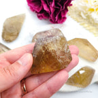 Hand holding a clear crystal with other crystals and flowers in the background. Citrine (Natural) Free Form for Abundance & Manifestation | Crystals Australia Inner Nurture