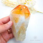 Hand holding a crystal with orange and white colors on a light background. Citrine crystal point available online at Inner Nurture Crystals Australia