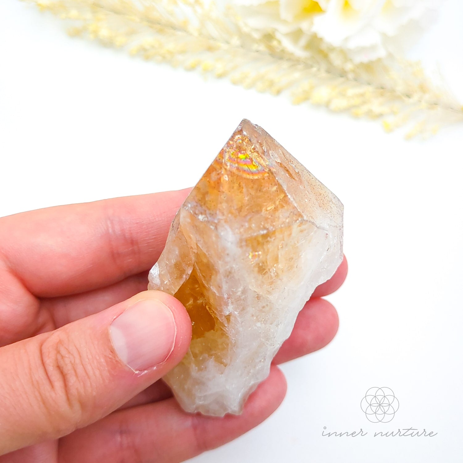 Hand holding a clear crystal with yellow interior, blurred flowers in the background. Citrine crystal available online at Inner Nurture Crystals Australia