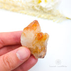Hand holding a clear crystal with orange inclusions against a white background. Citrine crystal available online at Inner Nurture Crystals Australia