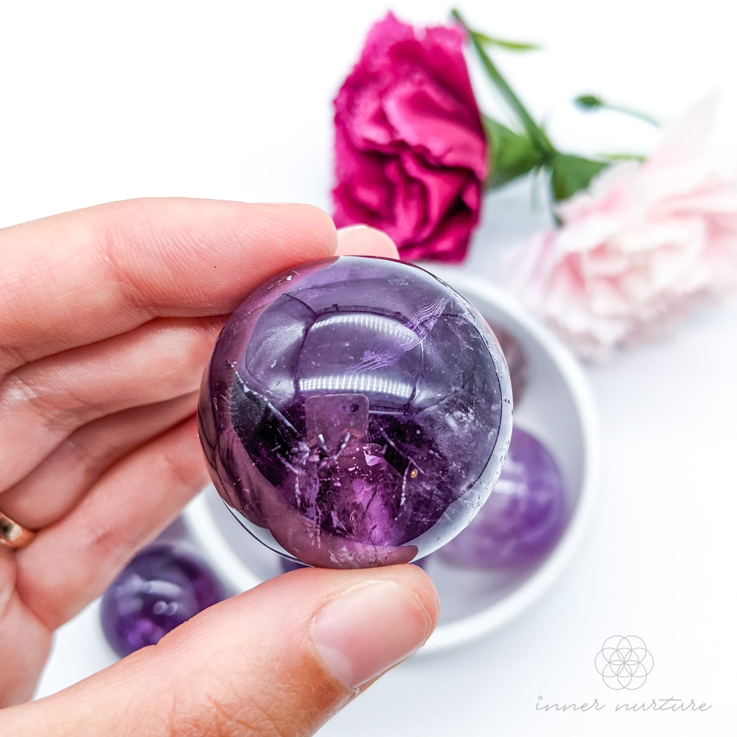 Hand holding a purple crystal ball with flowers in the background. Amethyst sphere available at Inner Nurture, an online crystal shop in Australia. 