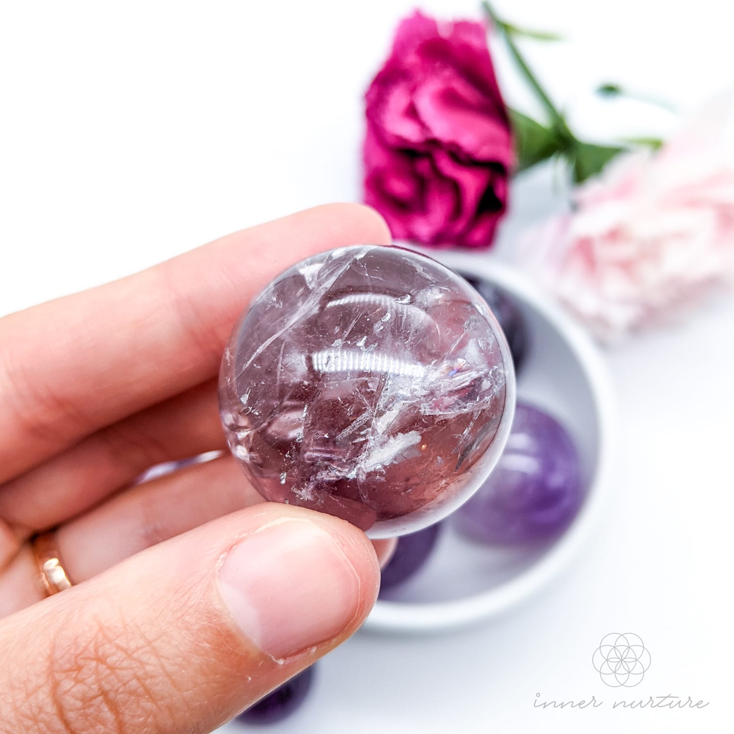 Hand holding a purple crystal ball with flowers in the background. Amethyst sphere available at Inner Nurture, an online crystal shop in Australia. 