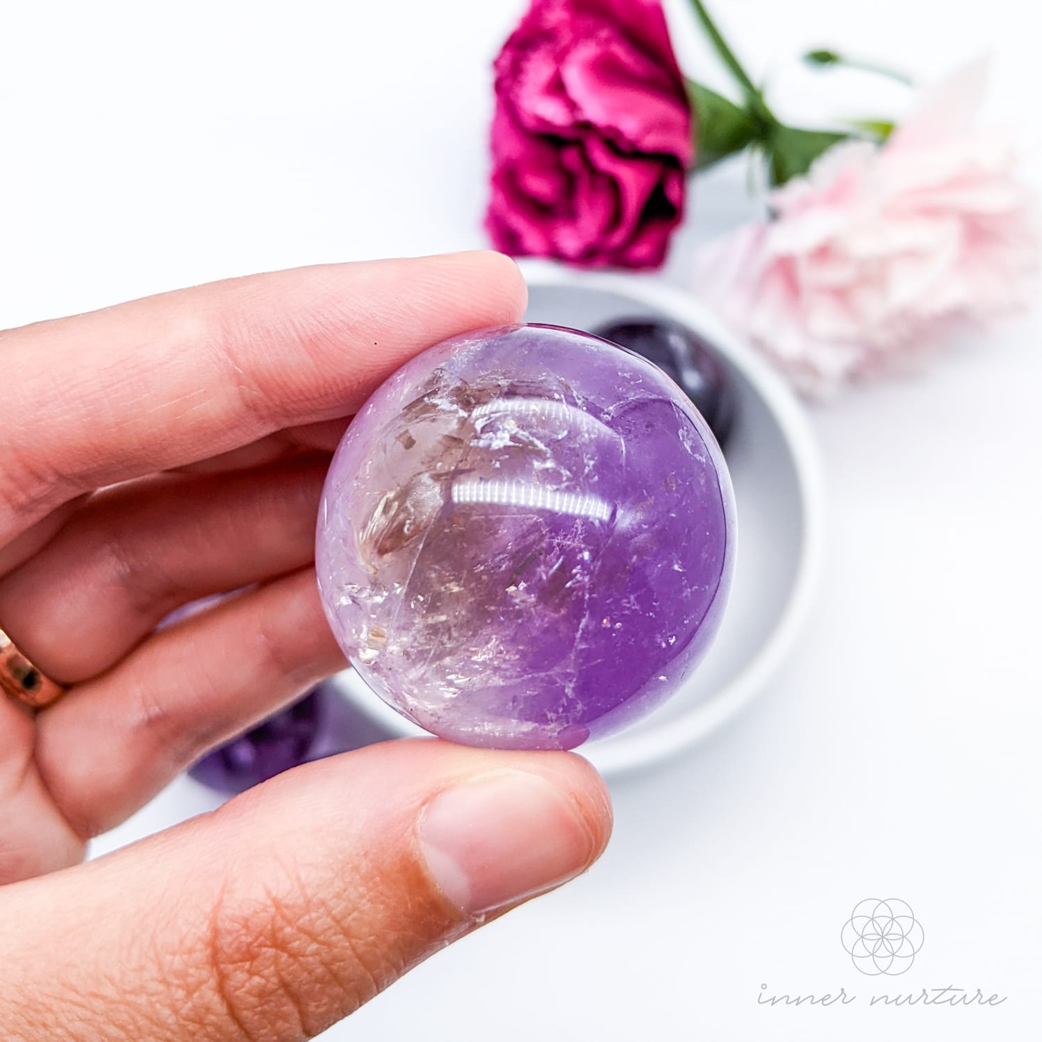 Hand holding a purple crystal ball with flowers in the background. Amethyst sphere available at Inner Nurture, an online crystal shop in Australia. 