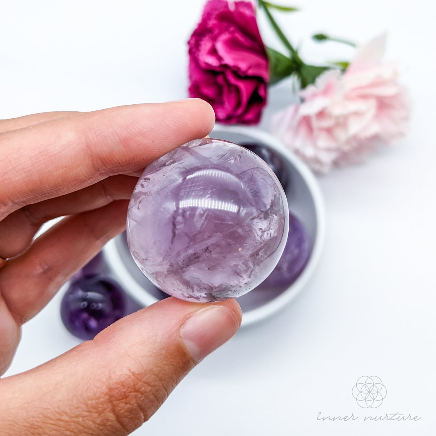 Hand holding a purple crystal ball with flowers in the background. Amethyst sphere available at Inner Nurture, an online crystal shop in Australia. 