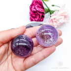Hand holding a purple crystal ball with flowers in the background. Amethyst sphere available at Inner Nurture, an online crystal shop in Australia. 